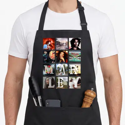Tablier de cuisine noir avec un motif en grille montrant Bob Marley, Trench Town et les lettres MAY sur fond tropical.