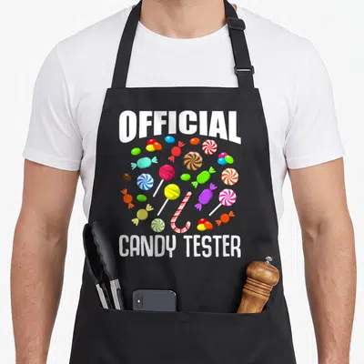 Tablier de cuisine noir avec l'inscription Official Candy Tester entouré de bonbons colorés et de sucreries variées.