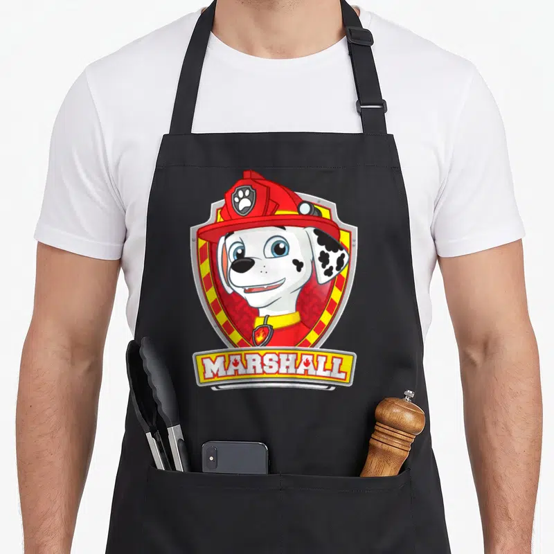 Tablier de cuisine noir avec l'illustration du chien pompier Marshall dans un insigne rouge et jaune portant son nom.