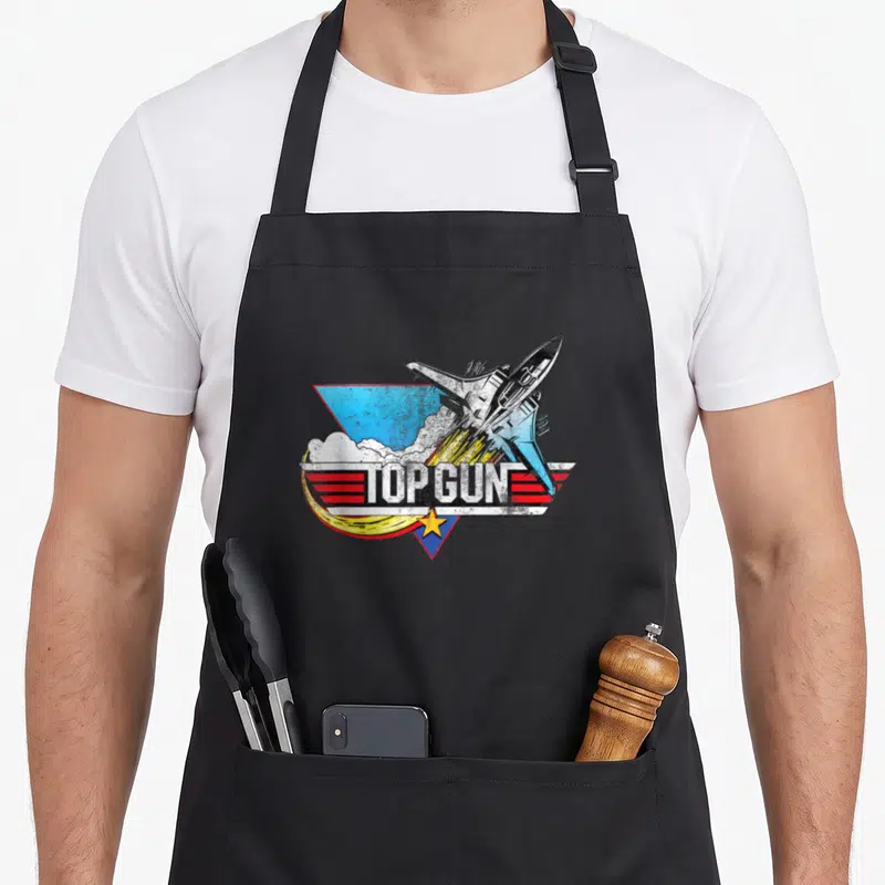 Tablier de cuisine noir avec un avion de chasse stylisé, le texte Top Gun, des ailes rouges et une étoile jaune sur fond géométrique.