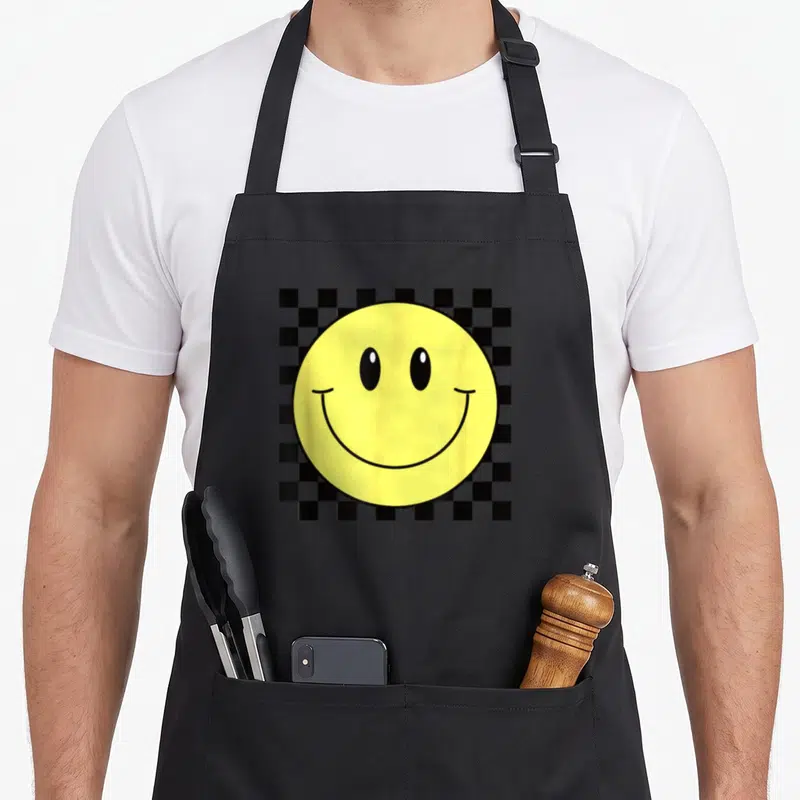Tablier de cuisine noir avec grand visage souriant jaune sur motif damier noir et blanc à l'effigie des années septante.