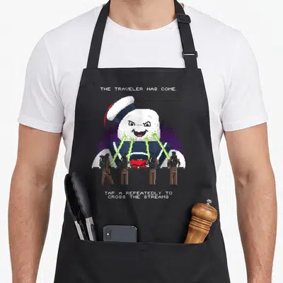 Tablier de cuisine noir avec illustration pixelisée du Stay Puft Marshmallow Man et personnages en tenues brownie tirant des rayons verts.