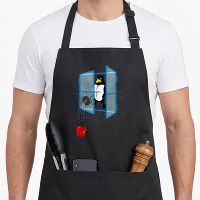 Tablier de cuisine noir avec un pingouin musclé brisant une fenêtre bleue et une pomme rouge mordue tombant en dessous.