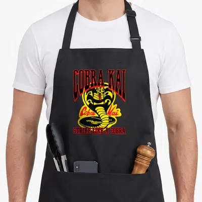 Tablier de cuisine noir avec l'emblème du cobra et les mots Cobra Kai Strike Like a Cobra imprimés en rouge et jaune.