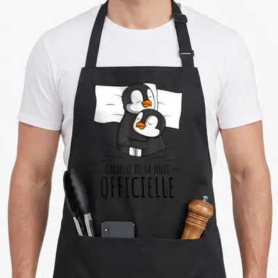 Tablier de cuisine noir avec deux pingouins s'enlaçant sous une couette et le texte « chemise de la nuit ».
