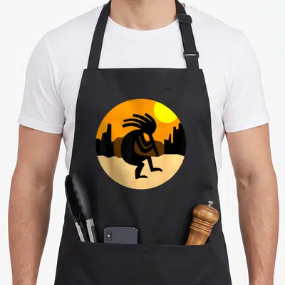Tablier de cuisine noir avec illustration circulaire d'un Kokopelli marchant devant un sunset orange et des silhouettes de cactus.
