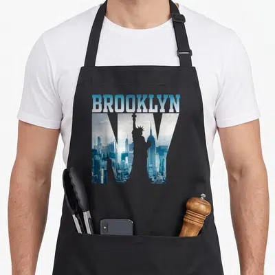 Tablier de cuisine noir avec linscription BROOKLYN et une silhouette de la Statue de la Liberté devant un paysage urbain bleu.