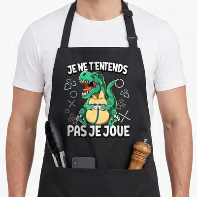 Tablier de cuisine noir avec un dinosaure vert tenant une manette et le texte Je ne t'entends pas je joue.