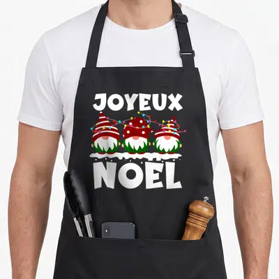 Tablier de cuisine noir avec trois gnomes de Noël aux chapeaux rouges et des guirlandes lumineuses colorées sur un fond blanc neigeux.