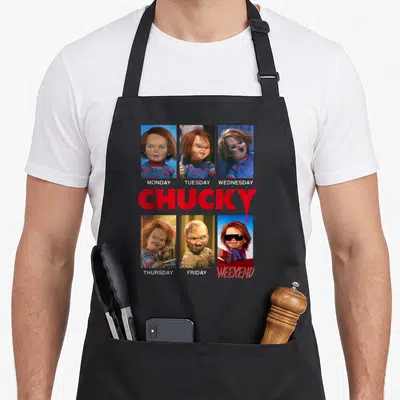 Tablier de cuisine noir avec six photos de Chucky représentant les jours de la semaine et le mot CHUCKY en rouge au centre.