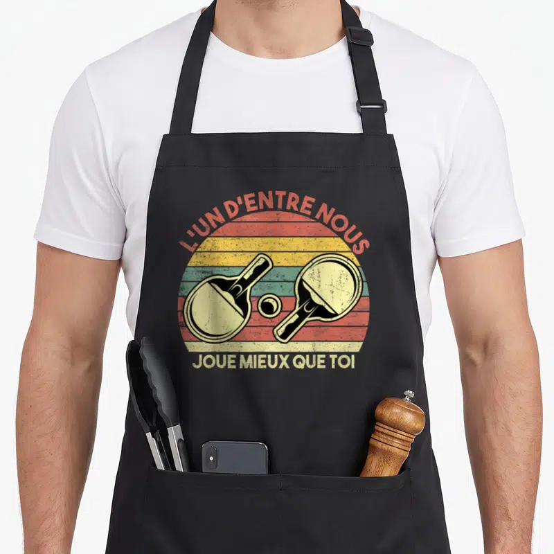Tablier de cuisine noir avec illustration vintage de deux raquettes de tennis de table et un ballon sur fond de soleil coloré.