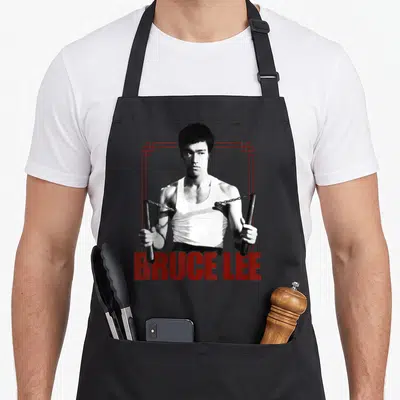 Tablier de cuisine noir avec illustration de Bruce Lee tenant des nunchaks et son nom en lettres rouges.