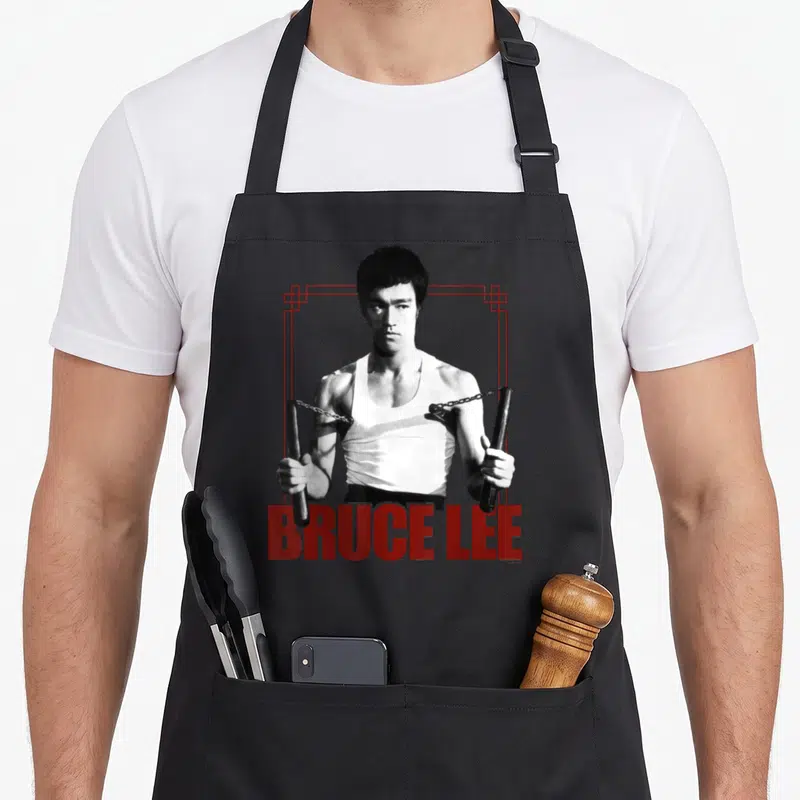 Tablier de cuisine noir avec illustration de Bruce Lee tenant des nunchaks et son nom en lettres rouges.