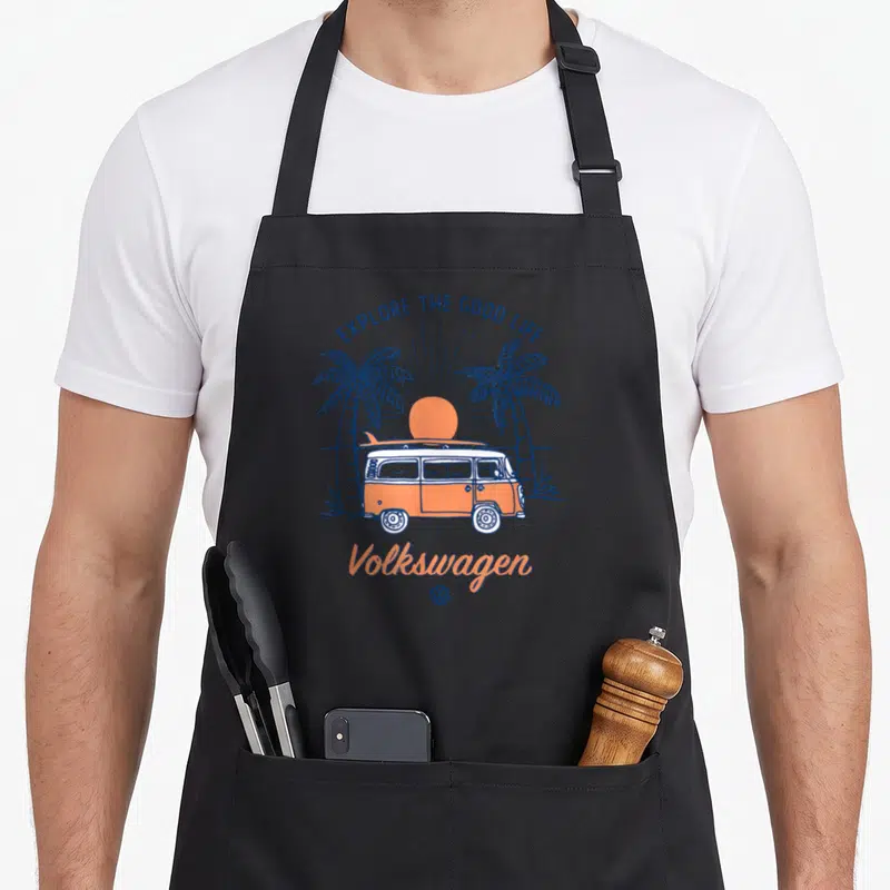 Tablier de cuisine noir avec illustration camping-car orange, palmiers et texte Volkswagen.
