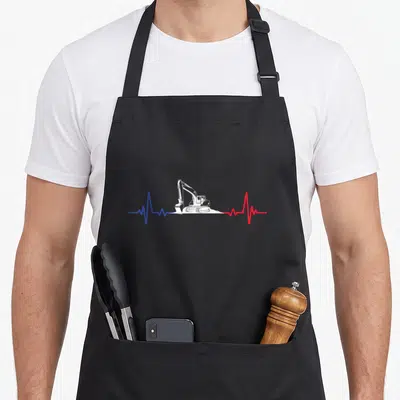 Tablier de cuisine noir avec une excavatrice blanche traversant une ligne de battement du cœur bleue et rouge sur fond uni.