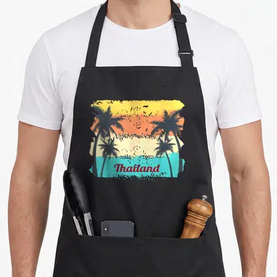 Tablier de cuisine noir avec illustration rétro de palmiers, ciel orange et turquoise, et le mot Thailand imprimé en bas.