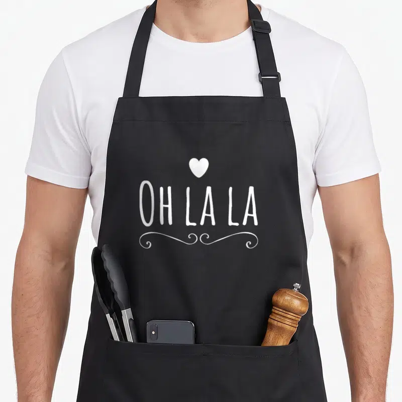 Tablier de cuisine noir avec inscription Oh la la, un cœur et des volutes décoratives en blanc au centre.