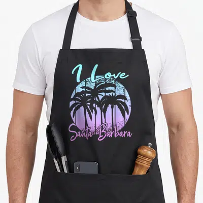 Tablier de cuisine noir avec un motif circulaire aux palmiers et texte J Love Santa Barbara en dégradé pastel.