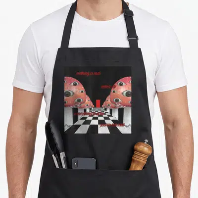 Tablier de cuisine noir avec des champignons roses à multiples yeux et texte rouge sur un sol en damier noir et blanc.