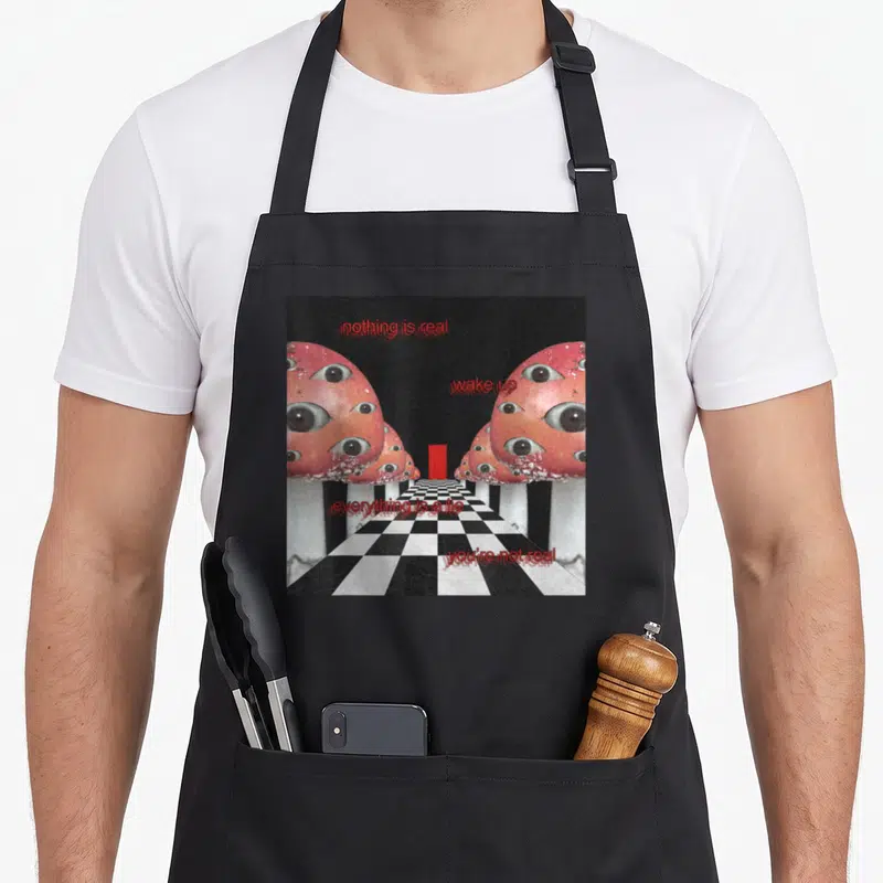 Tablier de cuisine noir avec des champignons roses à multiples yeux et texte rouge sur un sol en damier noir et blanc.