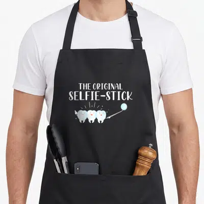 Tablier de cuisine noir avec trois dents souriantes tenant un miroir dentaire et le texte The Original Selfie-Stick.