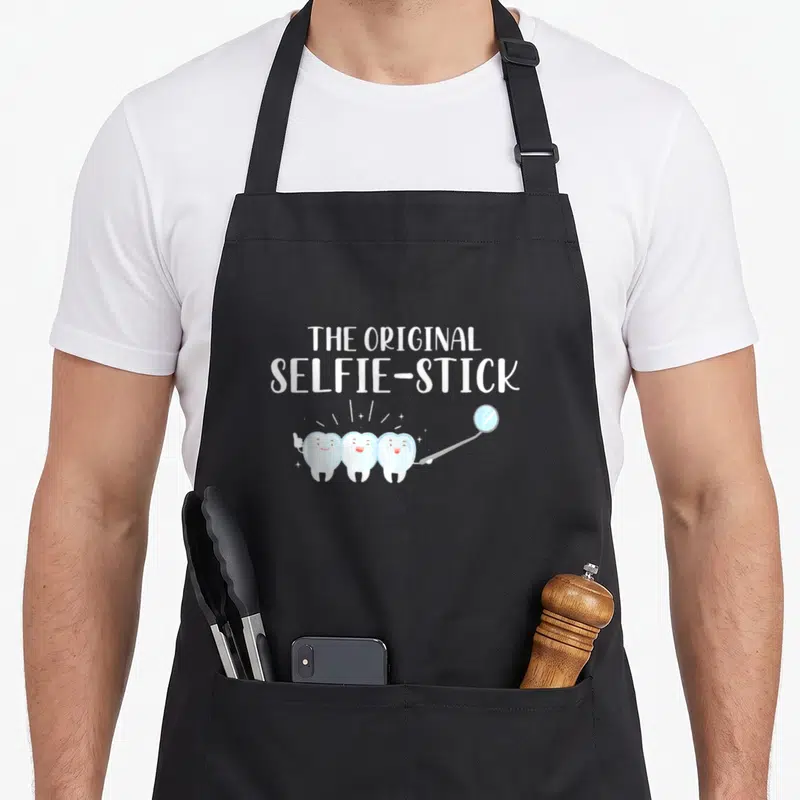 Tablier de cuisine noir avec trois dents souriantes tenant un miroir dentaire et le texte The Original Selfie-Stick.