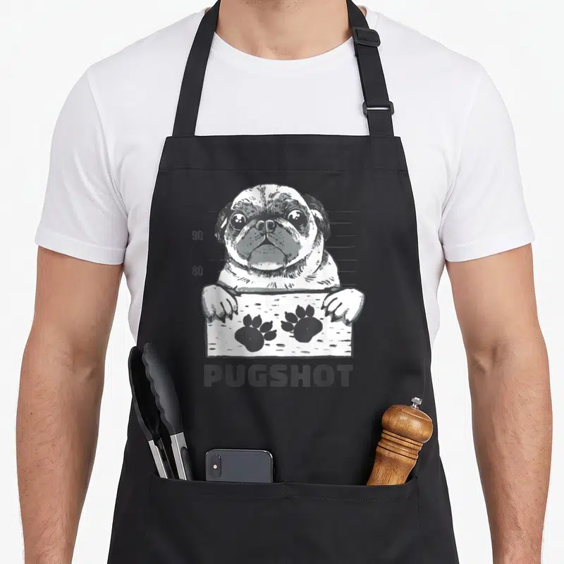 Tablier de cuisine noir avec illustration grise d'un pug devant un panneau d'empreintes de pattes et le mot Pugshot.