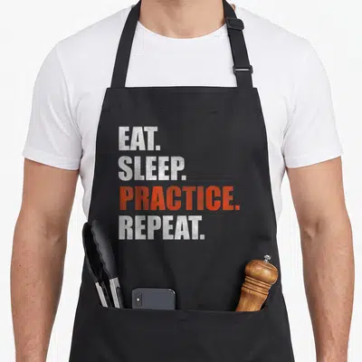 Tablier de cuisine noir avec le texte Eat Sleep Practice Repeat en lettres blanches et oranges disposées verticalement.