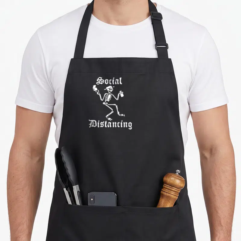 Tablier de cuisine noir avec un squelette blanc dansant tenant deux bouteilles et le texte Social Distancing en lettres gothiques.