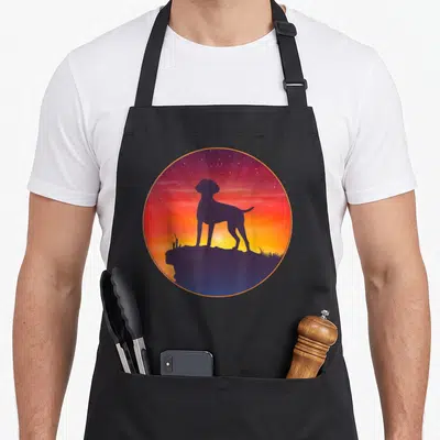 Tablier de cuisine noir avec une illustration circulaire représentant un chien en silhouette sur un rocher face à un ciel étoilé coloré.