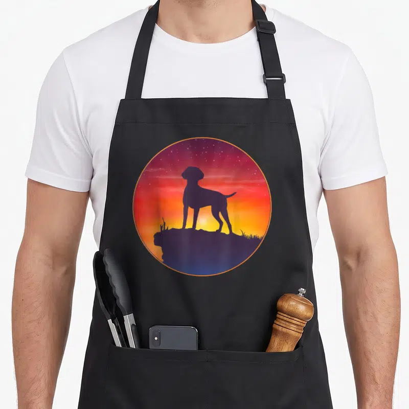 Tablier de cuisine noir avec une illustration circulaire représentant un chien en silhouette sur un rocher face à un ciel étoilé coloré.