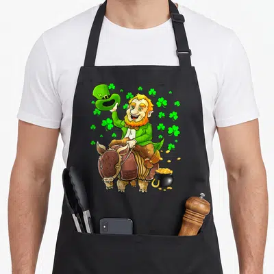 Tablier de cuisine noir avec illustration colorée d'un Leprechaun chevauchant un Armadillo entouré de trèfles verts et d'un pot de pièces d'or.