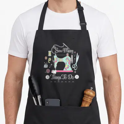 Tablier de cuisine noir avec une machine à coudre pastel, des bobines de fil et l'inscription Sew Many Things To Do en blanc.