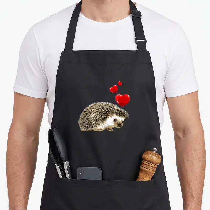 Tablier de cuisine noir avec illustration d'un hérisson aux piquants bruns et blancs, surmonté de trois cœurs rouges brillants flottant vers la droite.