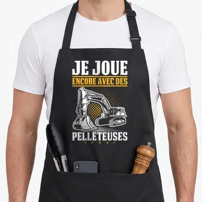 Tablier de cuisine noir avec une illustration de pelleteuse blanche et le texte Je joue encore avec des pelleteuses en blanc et jaune.
