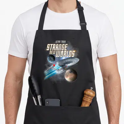 Tablier de cuisine noir avec illustration du vaisseau Enterprise, deux planètes et le titre Strange New Worlds en lettres dorées.