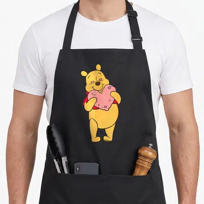 Tablier de cuisine noir avec Winnie l'ourson jaune tenant un cœur rose et des yeux en forme de petits cœurs roses.