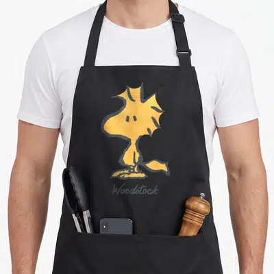 Tablier de cuisine noir avec l'illustration surdimensionnée de Woodstock, petit oiseau jaune au bec pointu et au nom écrit en dessous.