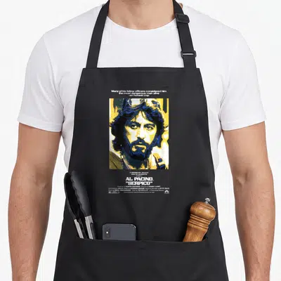 Tablier de cuisine noir avec une illustration jaune et bleue d'Al Pacino dans Serpico et du texte blanc en haut et en bas.