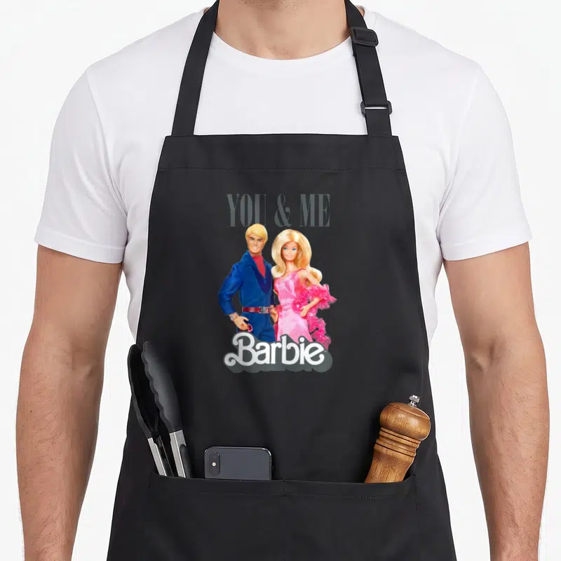 Tablier de cuisine noir avec Barbie en robe rose et Ken en costume bleu sous l'inscription You & Me.