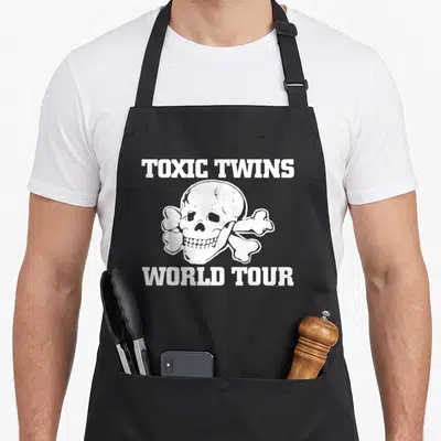 Tablier de cuisine noir avec un crâne blanc, des os croisés et le texte Toxic Twins World Tour imprimé en lettres capitales.