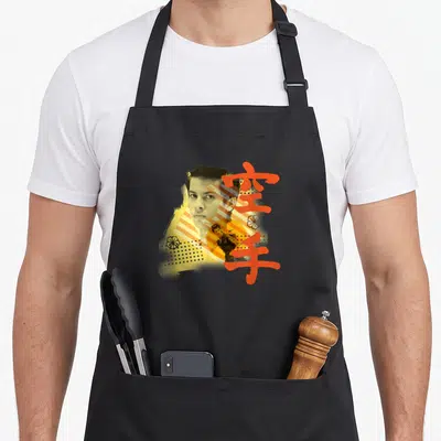 Tablier de cuisine noir avec portrait stylisé de Miguel, kanjis rouges et motifs géométriques orange et jaunes superposés.