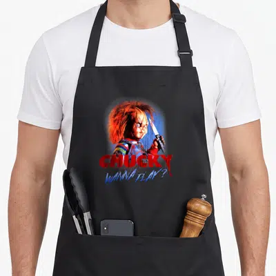 Tablier de cuisine noir avec l'illustration de Chucky tenant un couteau et le texte CHUCKY WANNA PLAY en rouge et bleu.