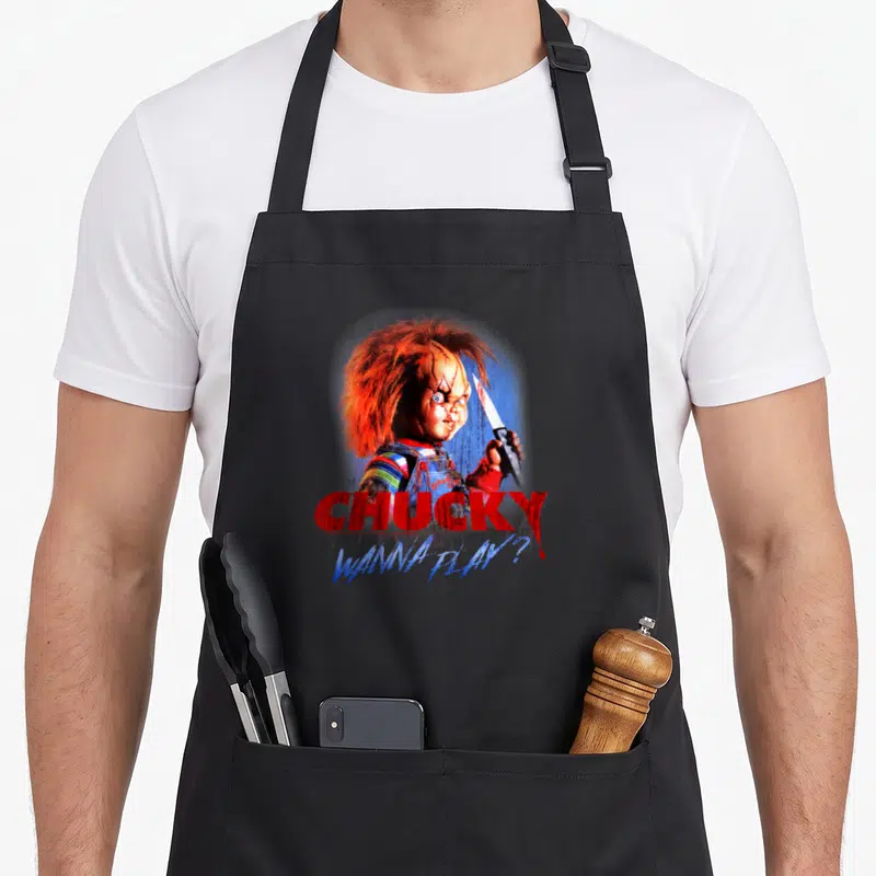 Tablier de cuisine noir avec l'illustration de Chucky tenant un couteau et le texte CHUCKY WANNA PLAY en rouge et bleu.