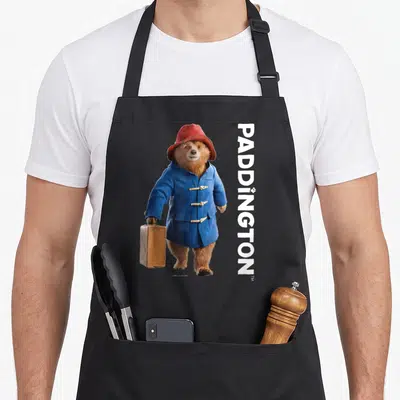 Tablier de cuisine noir avec Paddington en manteau bleu portant une valise et le mot PADDINGTON écrit verticalement.