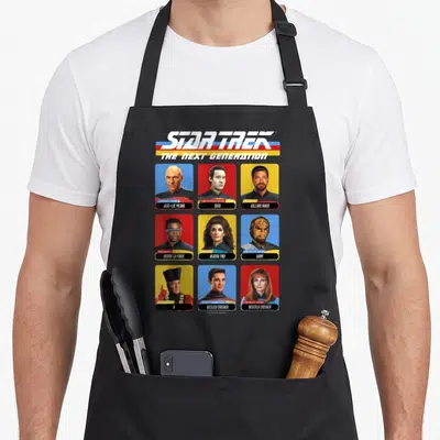 Tablier de cuisine noir avec les portraits stylisés des personnages de Star Trek La Nouvelle Génération sur fond coloré.