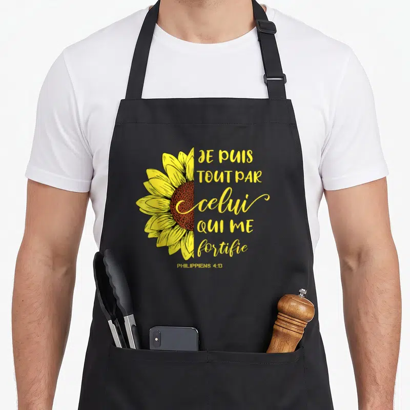 Tablier de cuisine noir avec un demi-tournesol jaune et le texte Je puis tout par celui qui me fortifie Philippiens 4:13 en lettres dorées.