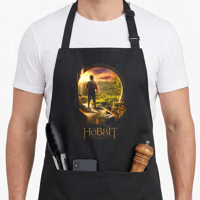Tablier de cuisine noir avec illustration de Bilbo Baggins regardant par une porte ronde vers un paysage verdoyant au coucher du soleil.