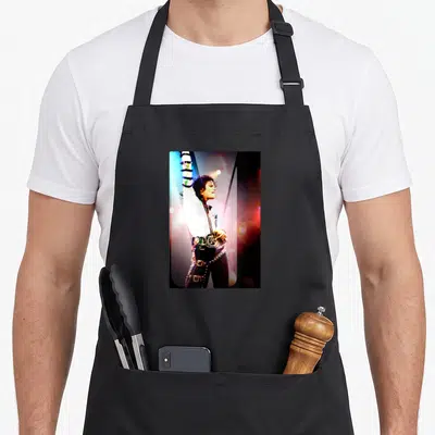 Tablier de cuisine noir avec une illustration colorée de Michael Jackson levant le bras sous des lumières de scène vives et contrastées.