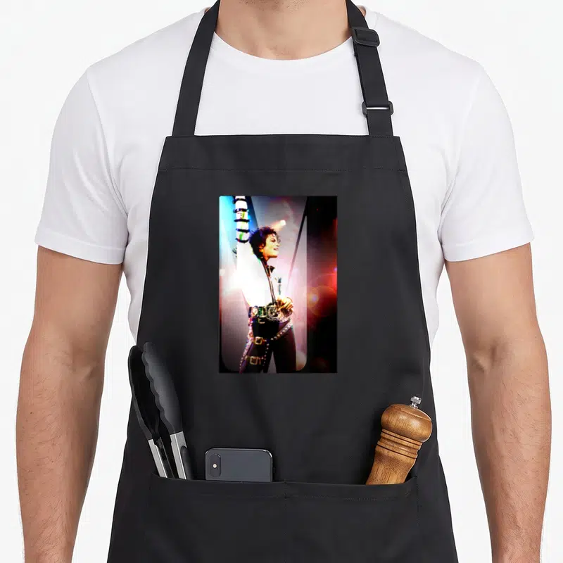 Tablier de cuisine noir avec une illustration colorée de Michael Jackson levant le bras sous des lumières de scène vives et contrastées.